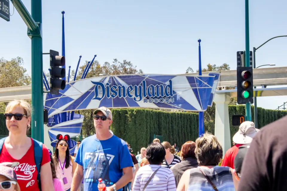 A crowd of people cross the street with a sign for the entrance of Disneyland in the background